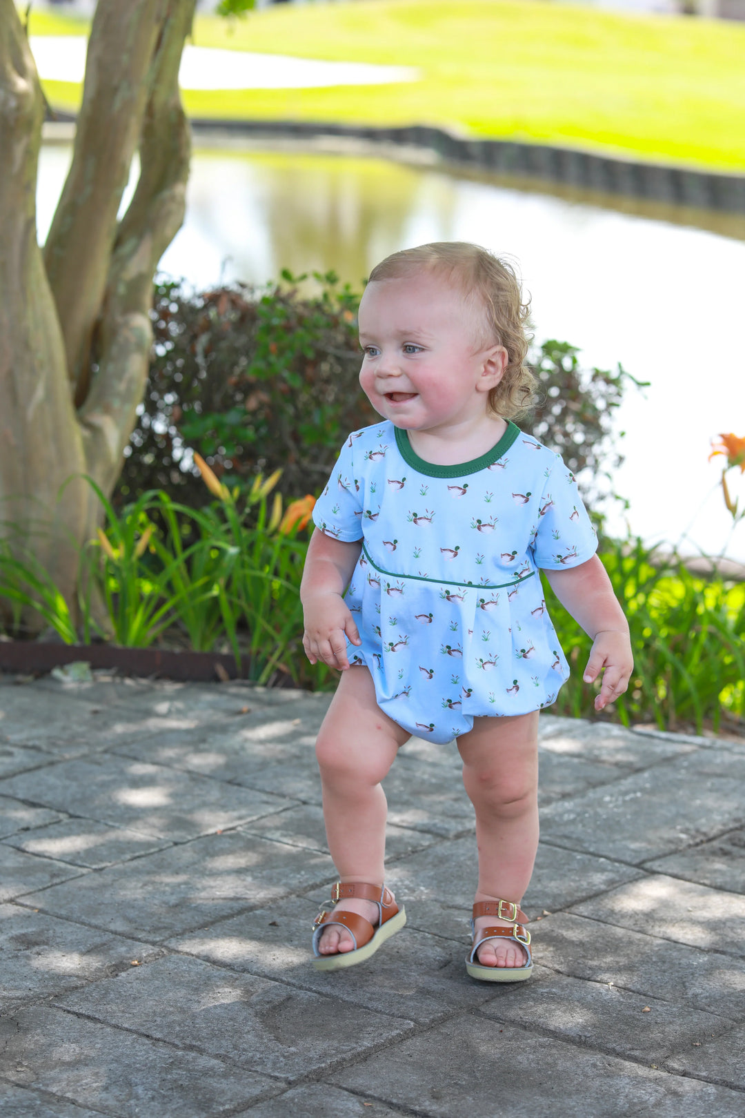 LITTLE BOY STANDING NEXT TO A POND WEARING BLUE OUTFIT WITH GREEN TRIM AND DUCK DESIGN