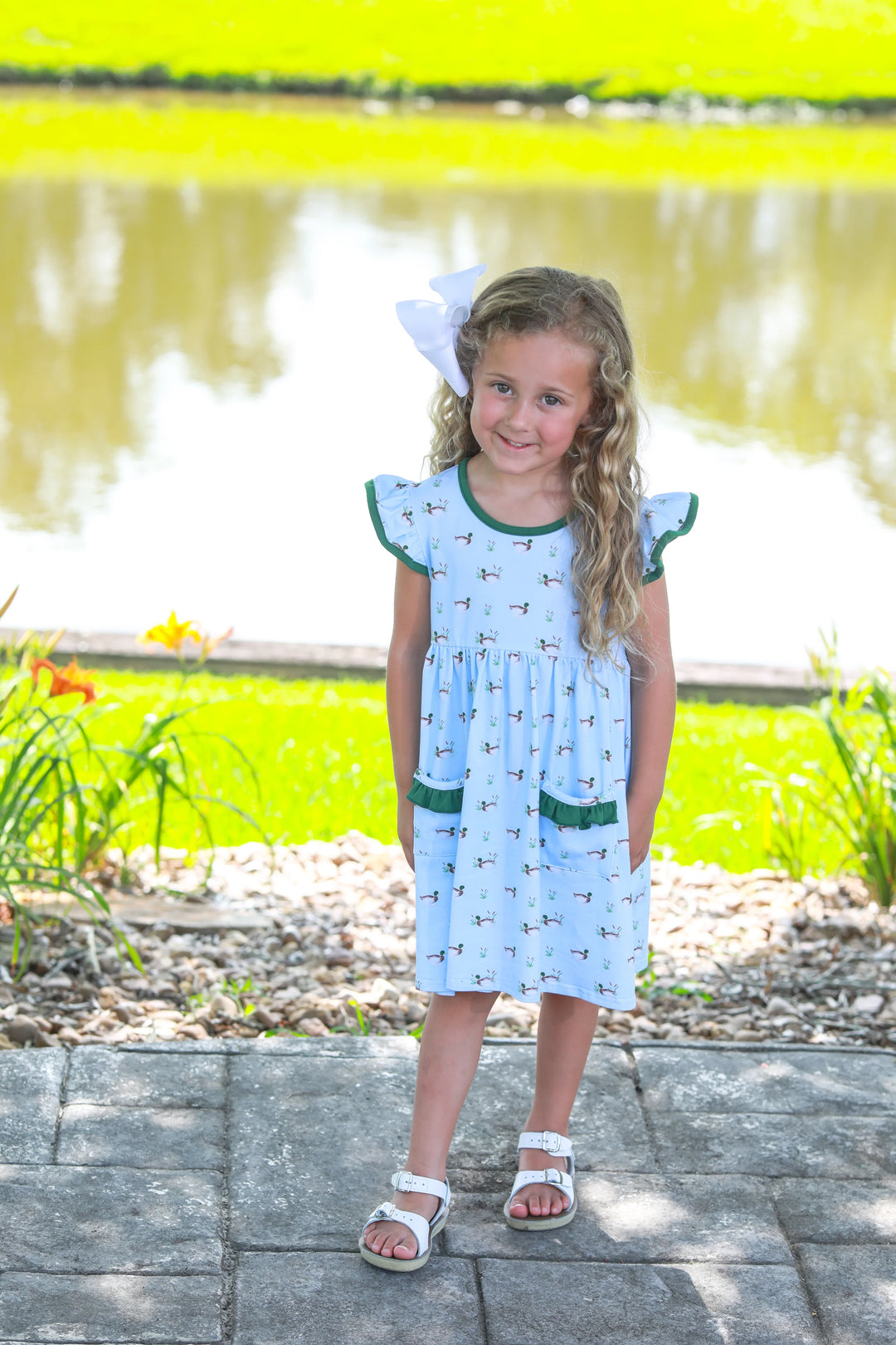 LITTLE GIRL WITH BLONDE HAIR STANDING NEXT TO POND WEARING BLUE DRESS WITH DUCKS AND GREEN RUFFLE TRIM