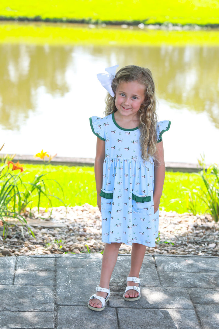 LITTLE GIRL WITH BLONDE HAIR STANDING NEXT TO POND WEARING BLUE DRESS WITH DUCKS AND GREEN RUFFLE TRIM