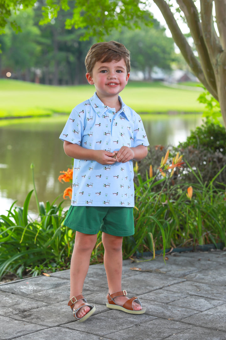 LITTLE BOY STANDING NEXT TO POND AND FLOWERS WEARING GREEN SHORTS AND BLUE COLLAR SHORT SLEEVE SHIRT WITH DUCKS ALL OVER