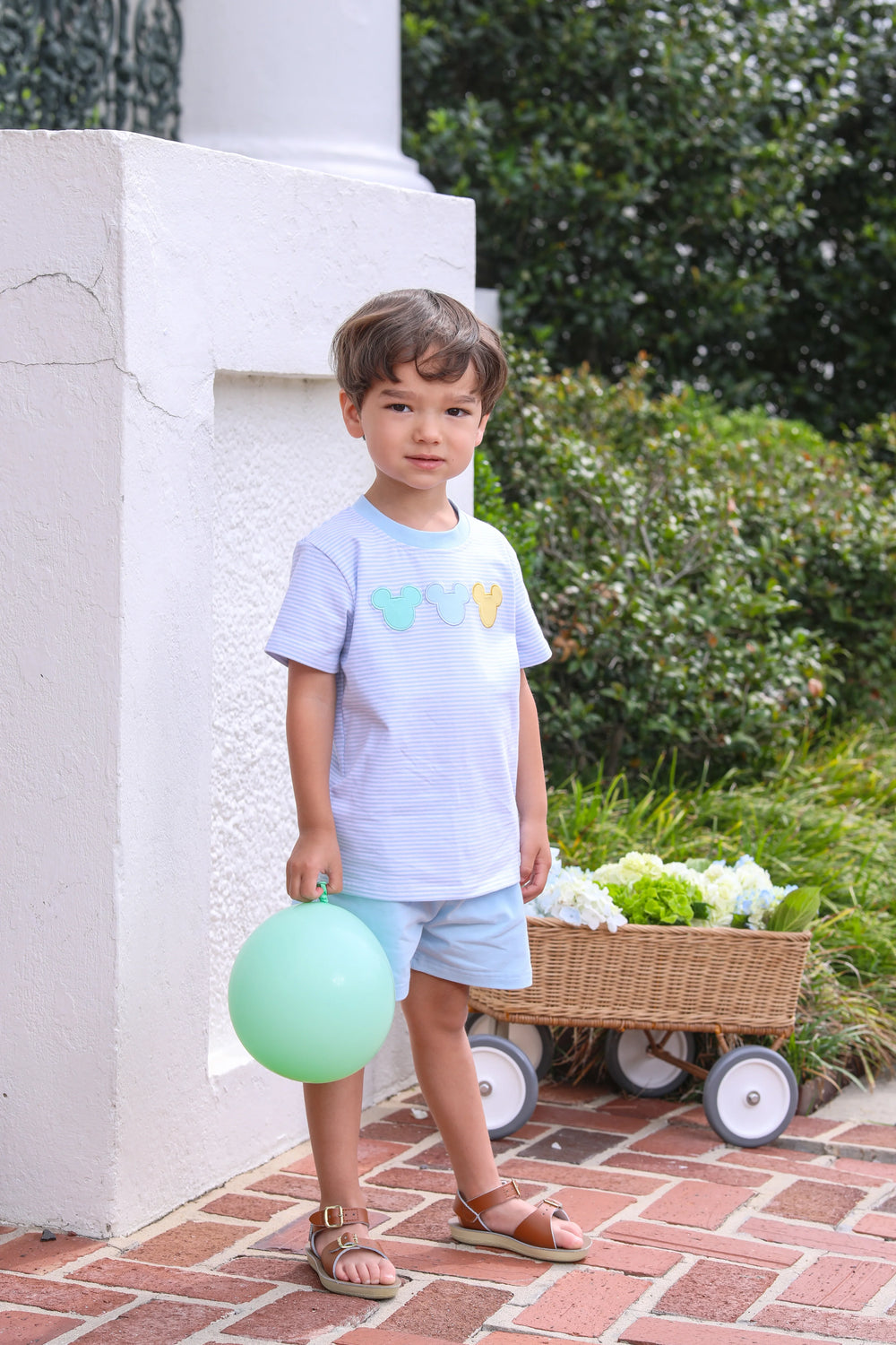 LITTLE BOY STANDING NEXT TO WHITE WALL WITH BALLOON WEARING BLUE AND WHITE STRIPE SHORT SET WITH DISNEY EARS