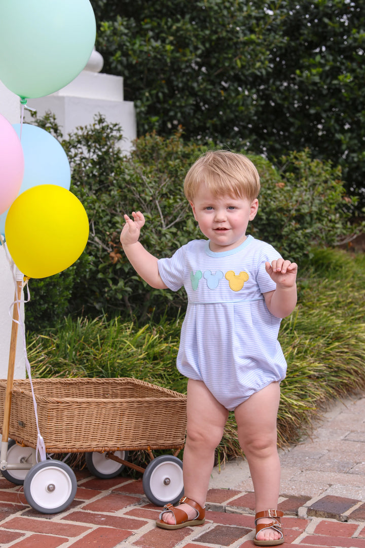 LITTLE BOY STANDING NEXT TO WICKER WAGON WITH BALLOONS WEARING BLUE STRIPED DISNEY EAR BUBBLE