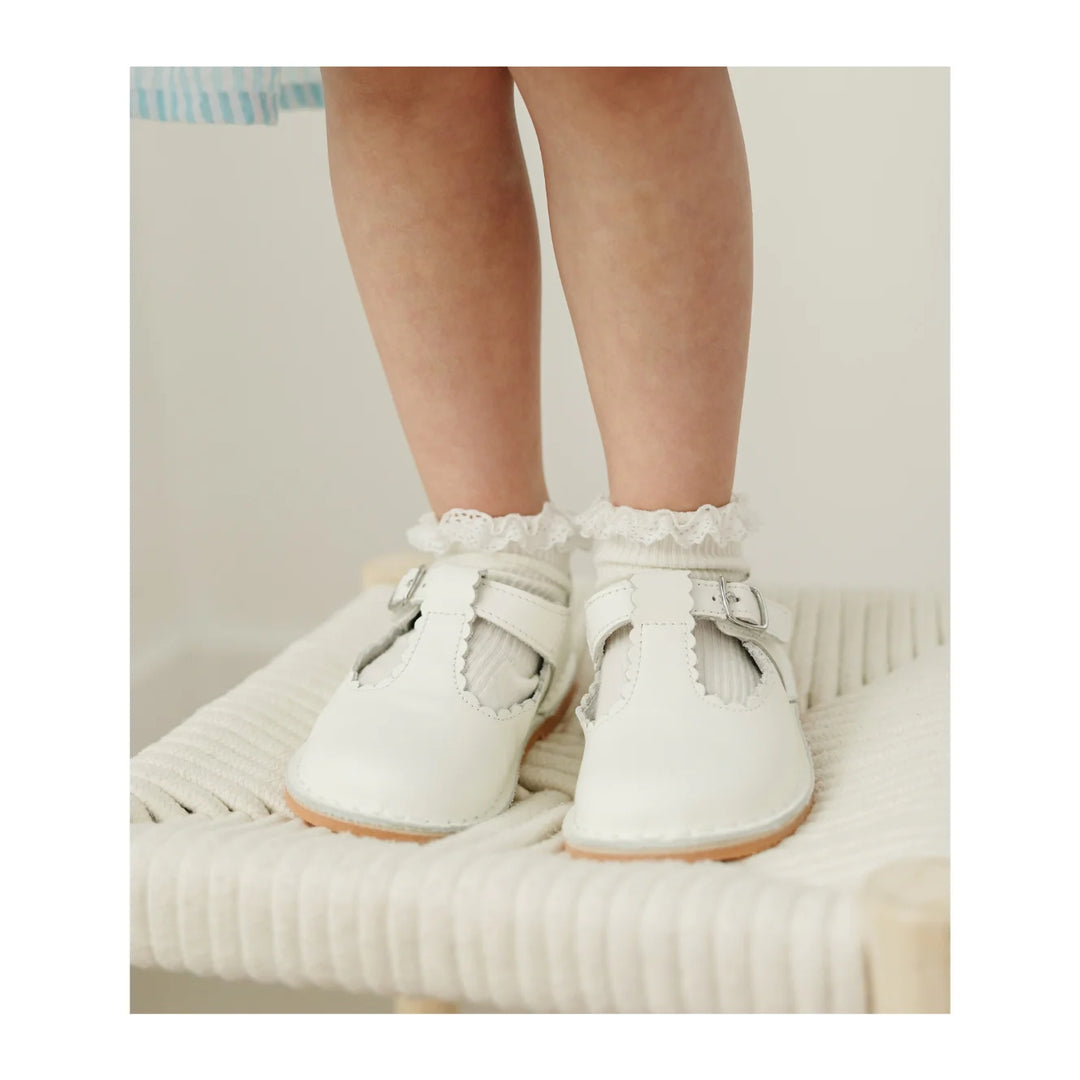 White baby shoes with ruffles worn by a child on a soft surface.