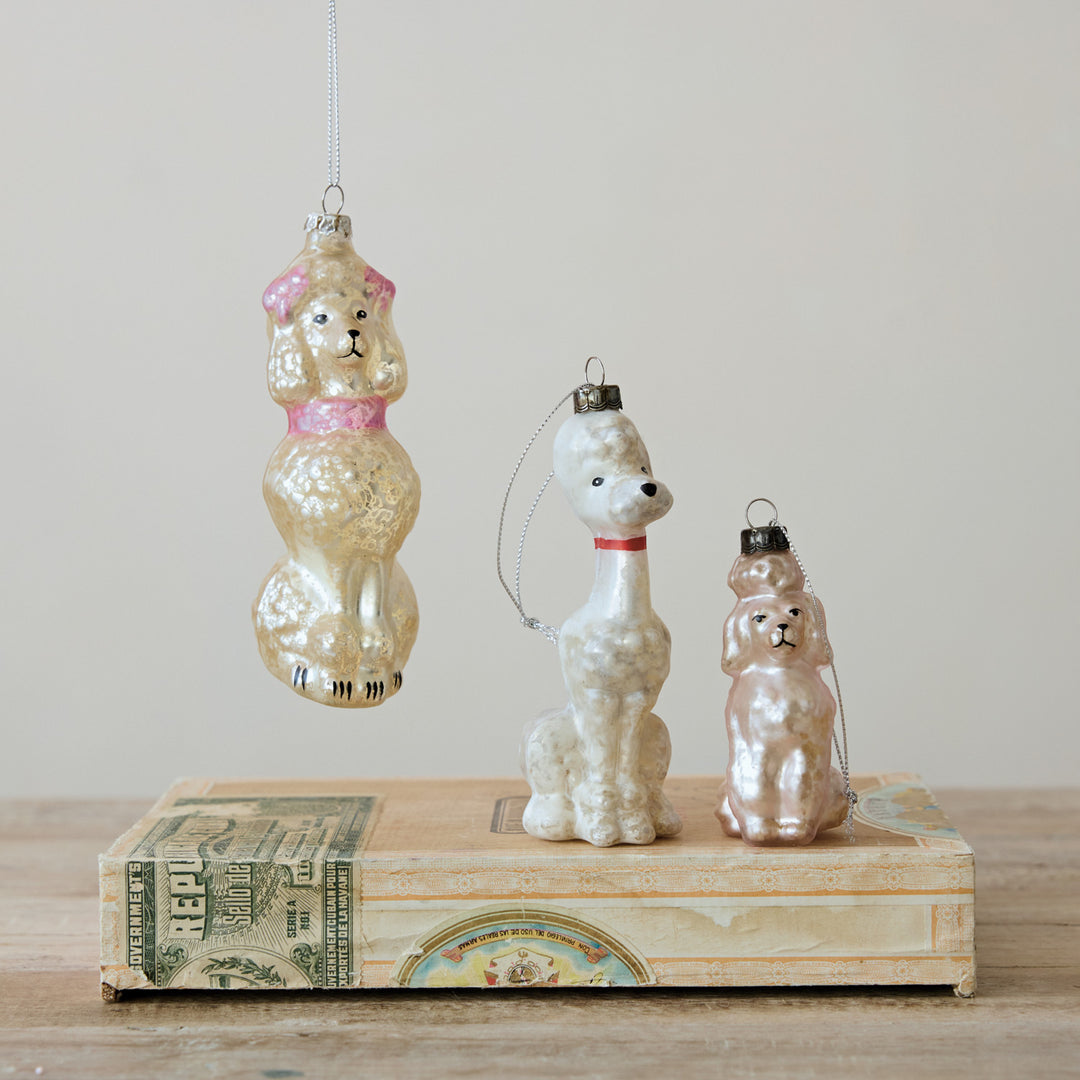 Three vintage poodle-shaped glass ornaments in gold, white, and rose gold tones are displayed on a stack of old dollar bills against a plain beige wall.