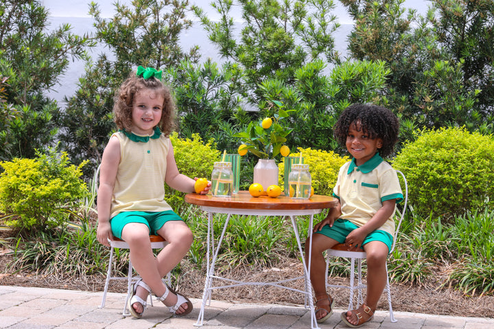 TWO LITTLE CHILDREN SITTING IN CHAIRS BY A TABLE WITH LEMONS AND DRINKS WEARING COORDINATING YELLOW AND GREEN OUTFITS