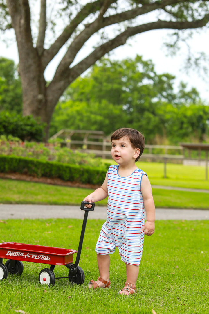 LITTLE BOY IN GRASS WEARING BLUE RED AND WHITE JON JON PULLING A WAGON