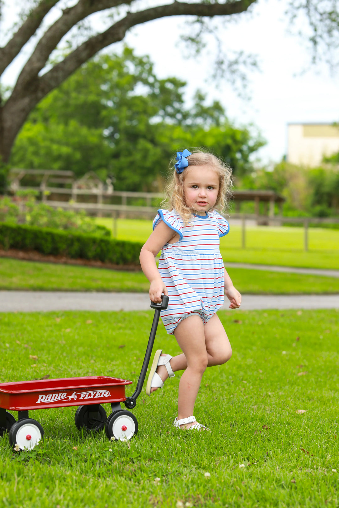 LITTLE GIRL IN THE GRASS PULLING A RED WAGON IN A BLUE RED AND WHITE FLUTTER SLEEVE PATRIOTIC BUBBLE