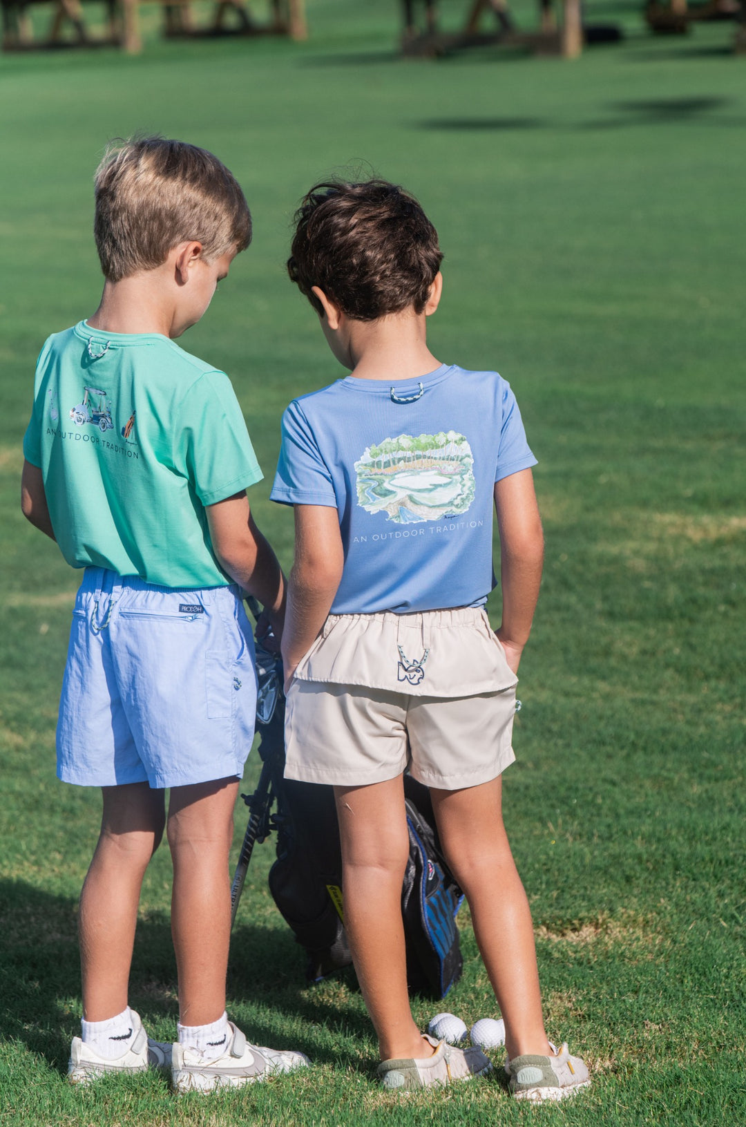 Two children standing on a grassy field wearing matching shirts and shorts.