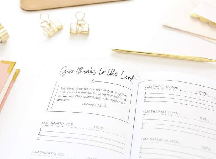 An open planner or journal spread on a white surface, featuring a handwritten note that says 'Give thanks to the Lord' with a Bible verse and lined sections for writing 'I AM THANKFUL FOR' and the date. Gold binder clips, a gold pen, and pastel paper accents are arranged around it.