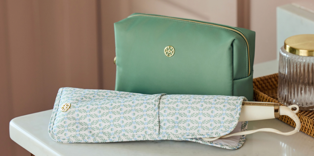 Two cosmetic pouches displayed on a tabletop: a larger sage green pouch with gold zipper and a circular gold logo, and a smaller light blue patterned pouch in front with a cream strap and gold logo. A glass jar and woven tray are partially visible in the background.