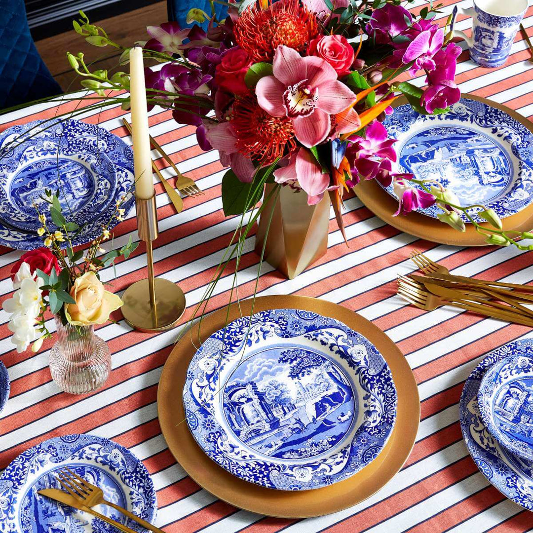 Table setting with blue and white plates, gold cutlery, and colorful flowers on a striped tablecloth.