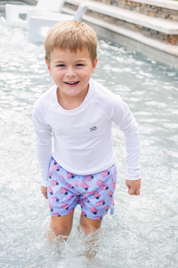 Young boy in a white long-sleeve shirt and patterned shorts standing in shallow water.