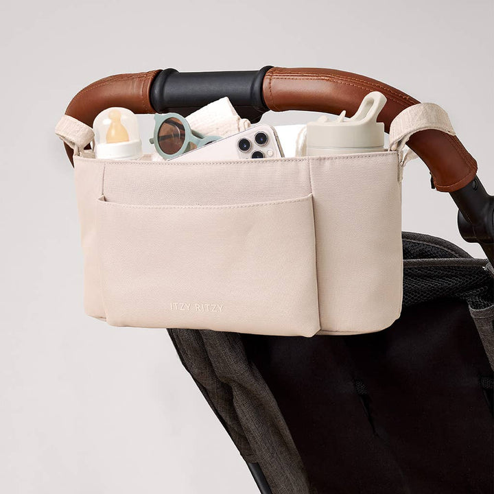 A light beige diaper bag organizer pouch mounted on a stroller handlebar, holding baby accessories like a bottle, sunglasses, and a phone. The stroller handlebar is brown with a black grip, set against a clean light gray background.