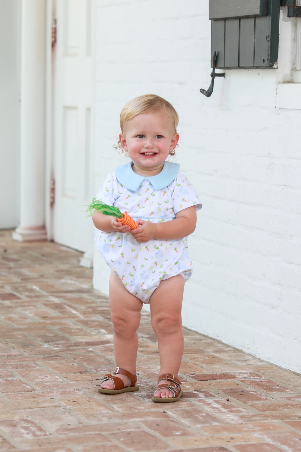 LITTLE BLOND BOY HOLDING CARROT STANDING NEXT TO WHITE WALL IN BLUE COLLAR WHITE BUBBLE WITH BUNNY AND CHICK DESIGN