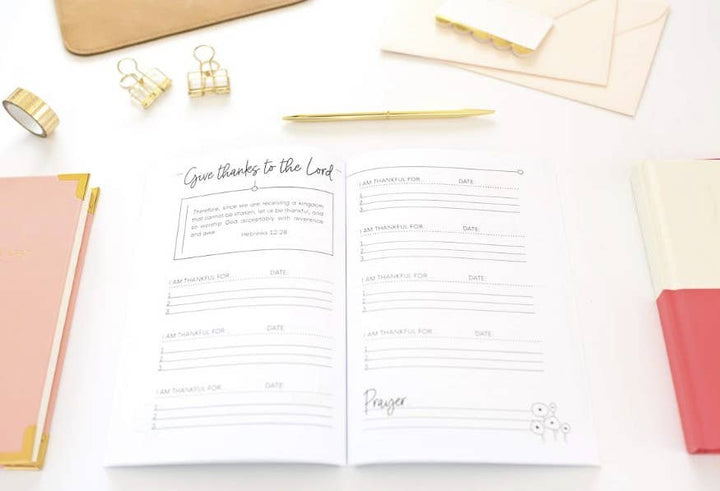 An open white planner or journal spread on a light surface, showing printed prayer and gratitude pages with lined sections and a small floral illustration. Surrounding props include a peach notebook, a coral-red notebook, gold binder clips, a gold pen, washi tape, and cream envelopes.