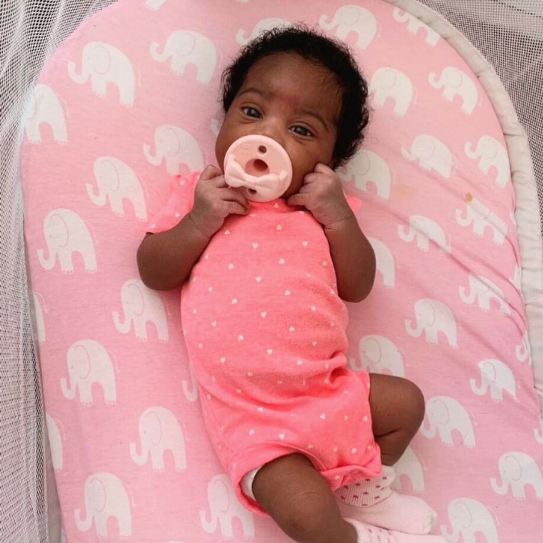 Baby in a pink outfit with a pacifier, lying on a pink pillow with elephant patterns.