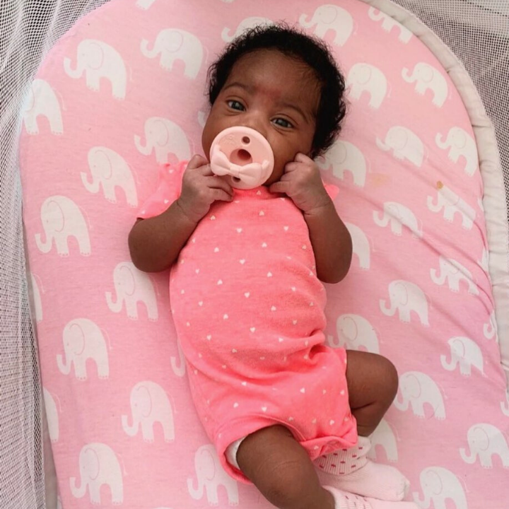 Baby in a pink outfit with a pacifier, lying on a pink pillow with elephant patterns.