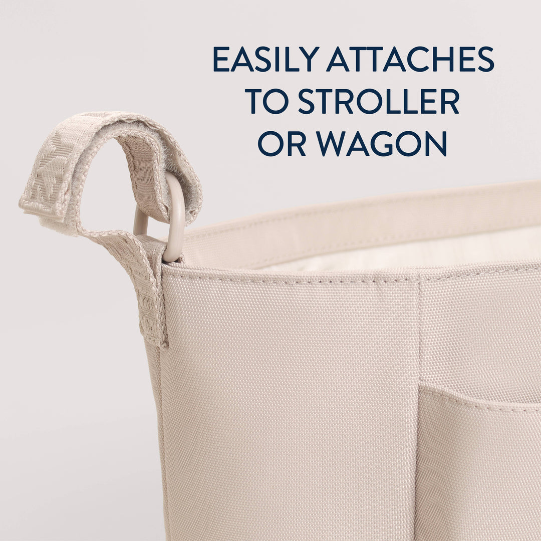 Close-up of a beige fabric storage basket with a loop handle, shown against a light gray background with dark blue text reading 'EASILY ATTACHES TO STROLLER OR WAGON'.