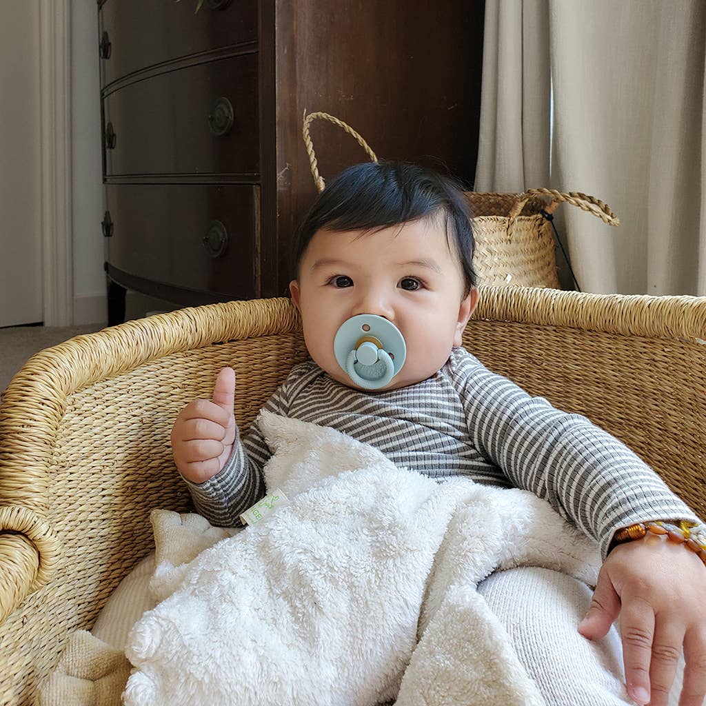 Baby sitting in a wicker chair with a pacifier, giving a thumbs-up gesture.