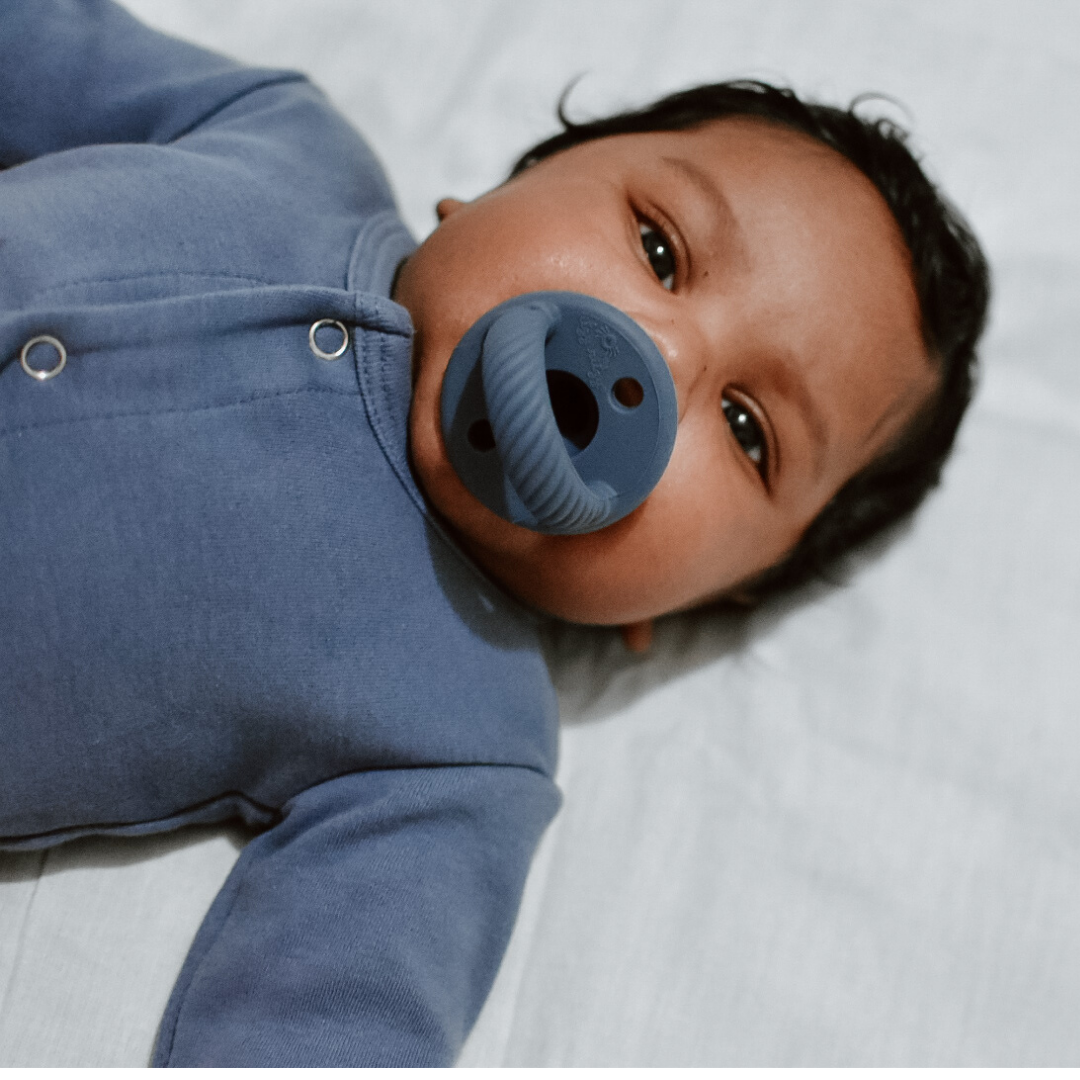 Baby wearing a blue outfit with a pacifier, lying on a white surface