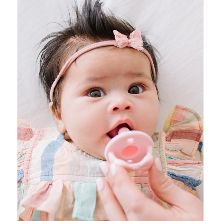 Baby with a pacifier held by an adult hand, wearing a colorful outfit and headband.