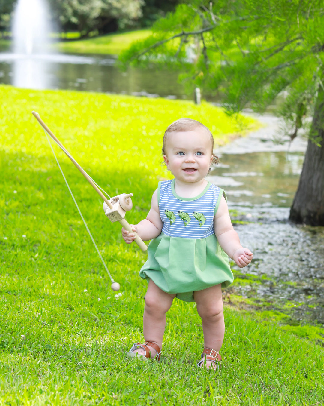 little boy in fishing trio bubble with green bottom and blue striped top. standing by pond holding fishing pole. 