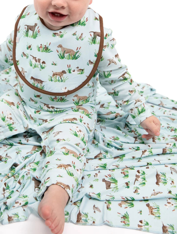 A baby wearing a light blue long-sleeve footed sleeper and matching bib, both printed with a pattern of brown dogs, green grass, and small wildflowers. The baby is sitting on a white background with the matching blanket spread out underneath.