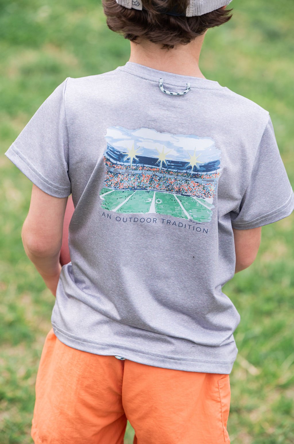 Child wearing a gray t-shirt with a graphic design on the back, standing in a grassy area.