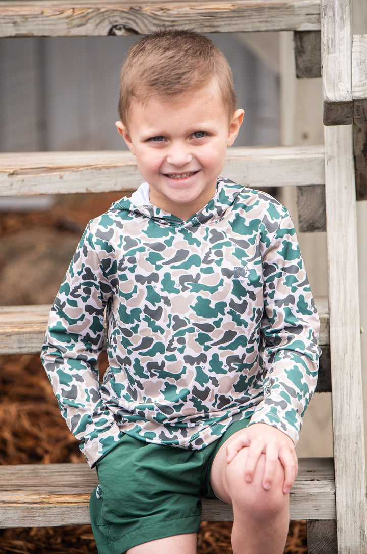 Young boy wearing a camouflage hoodie and green shorts sitting on wooden steps.