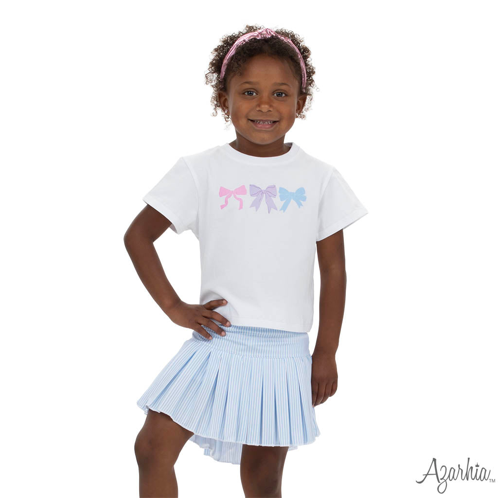 Young girl wearing a white t-shirt with colorful bow design and a light blue pleated skirt on a white background.