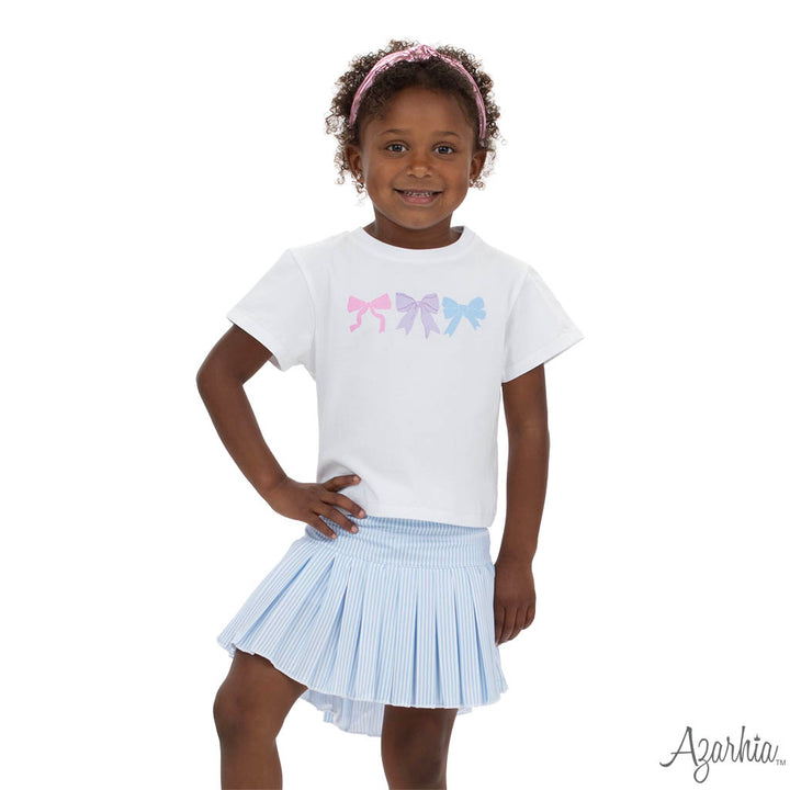 Young girl wearing a white t-shirt with colorful bow design and a light blue pleated skirt on a white background.