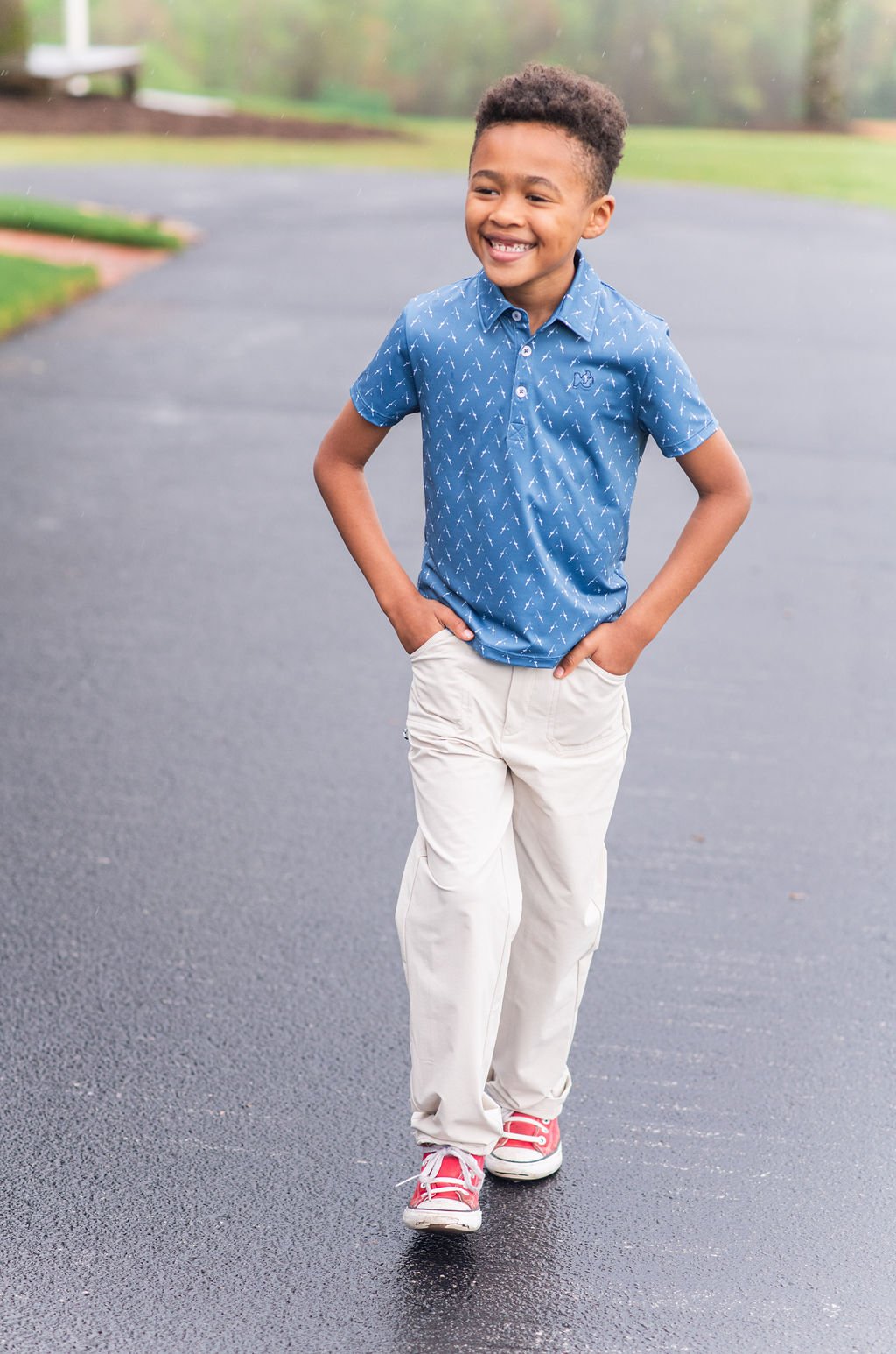Child in a blue polo shirt and white pants standing on a paved road.