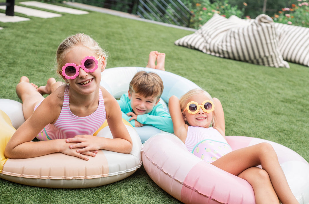 Children lying on inflatable pool toys with sunglasses on a grassy area.