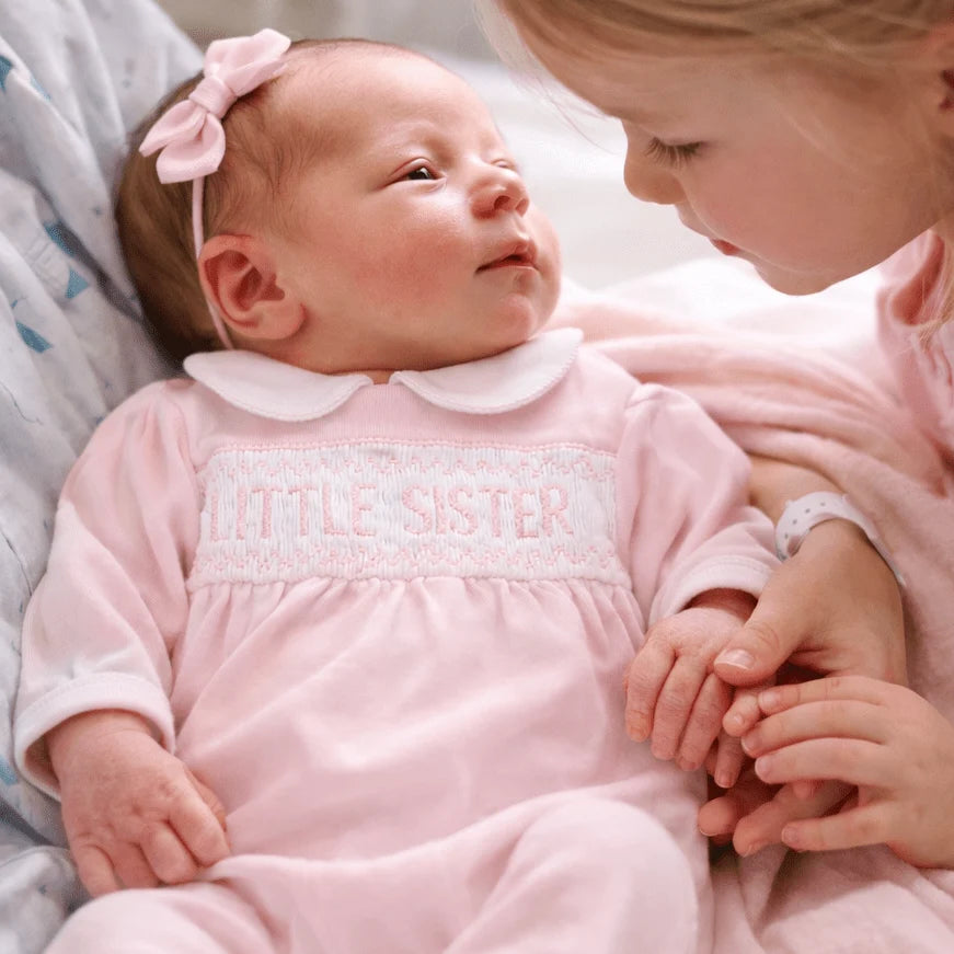 Newborn baby in a pink 'Little Sister' outfit held by an older child, both wearing matching pink outfits.