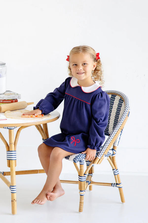 Young girl in a navy dress with a red bow sitting on a chair with striped cushions.