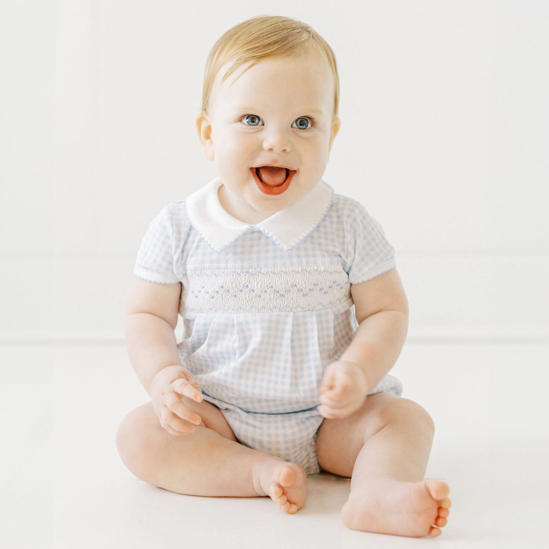Baby sitting on a white surface wearing a checkered outfit with a white collar.
