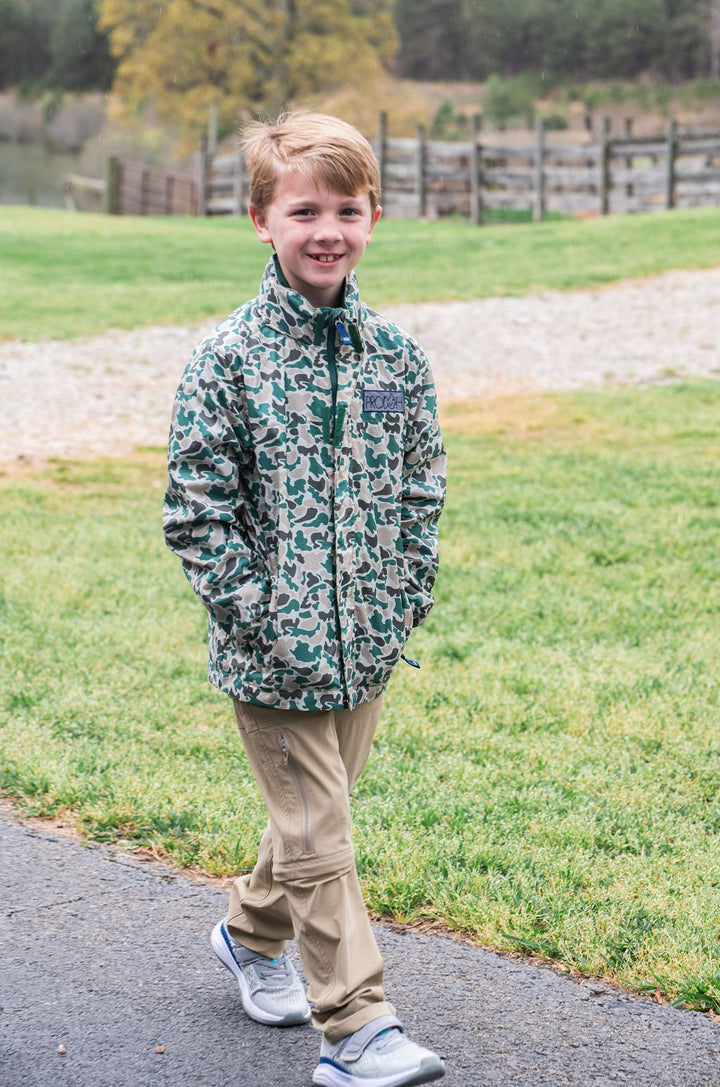 Young boy wearing a green camouflage jacket standing on a path with grass and trees in the background