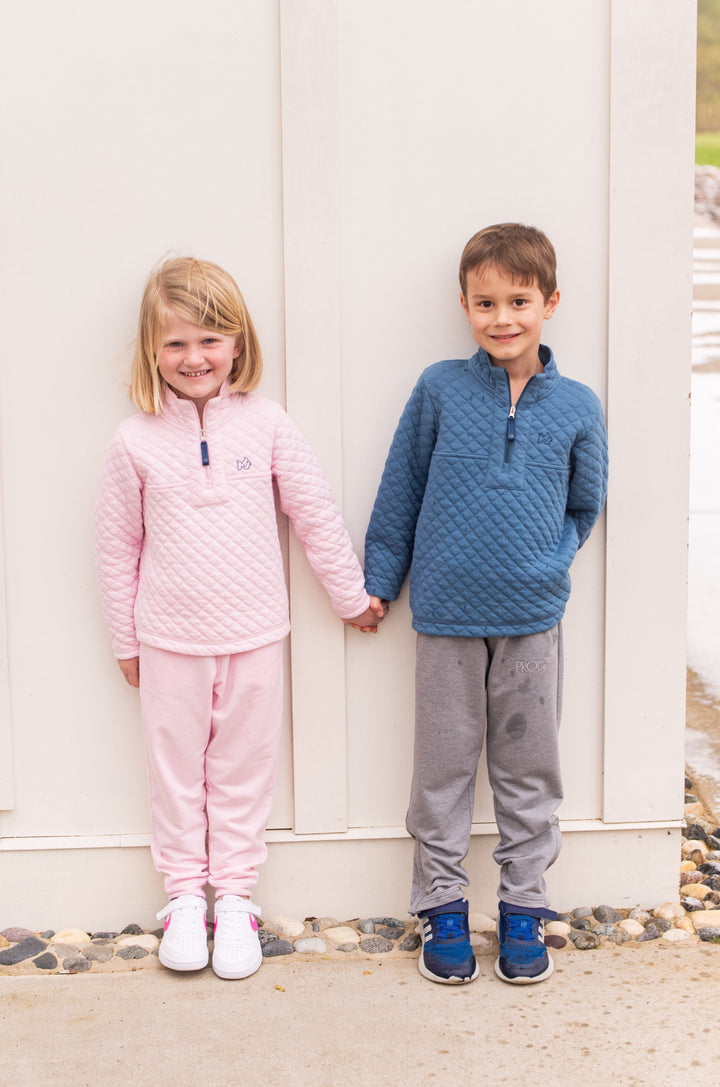 Two children in matching quilted jackets standing outdoors against a white wall.