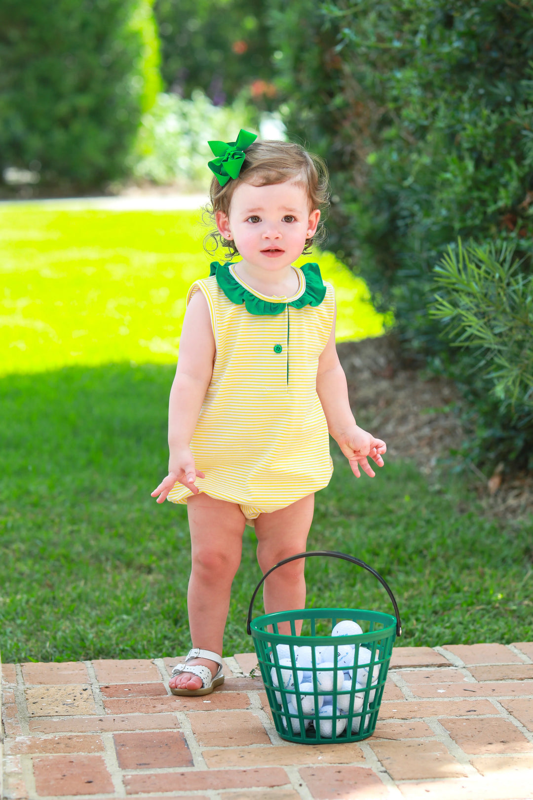 LITTLE GIRL WITH GREEN BOW IN HER HAIR STANDING NEXT TO BUCKET OF GOLF BALLS IN YELLOW STRIPED BUBBLE WITH GREEN RUFFLE TRIM AROUND COLLAR