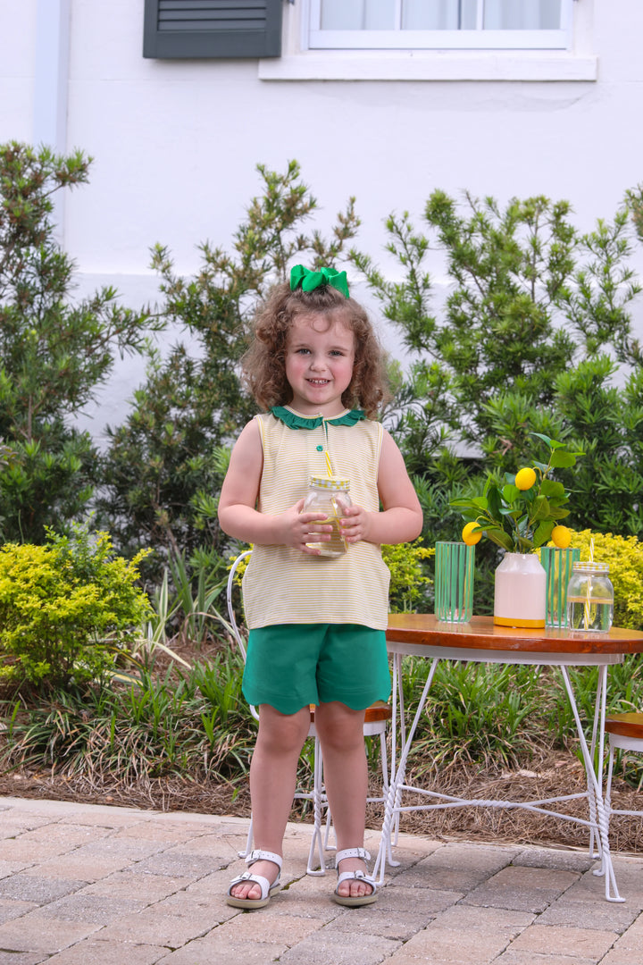 LITTLE GIRL HOLDING DRINK WEARNG YELLOW STRIPED TANK TOP WITH GREEN RUFFLE TRIM AND MATCHING GREEN SCALLOPED SHORTS