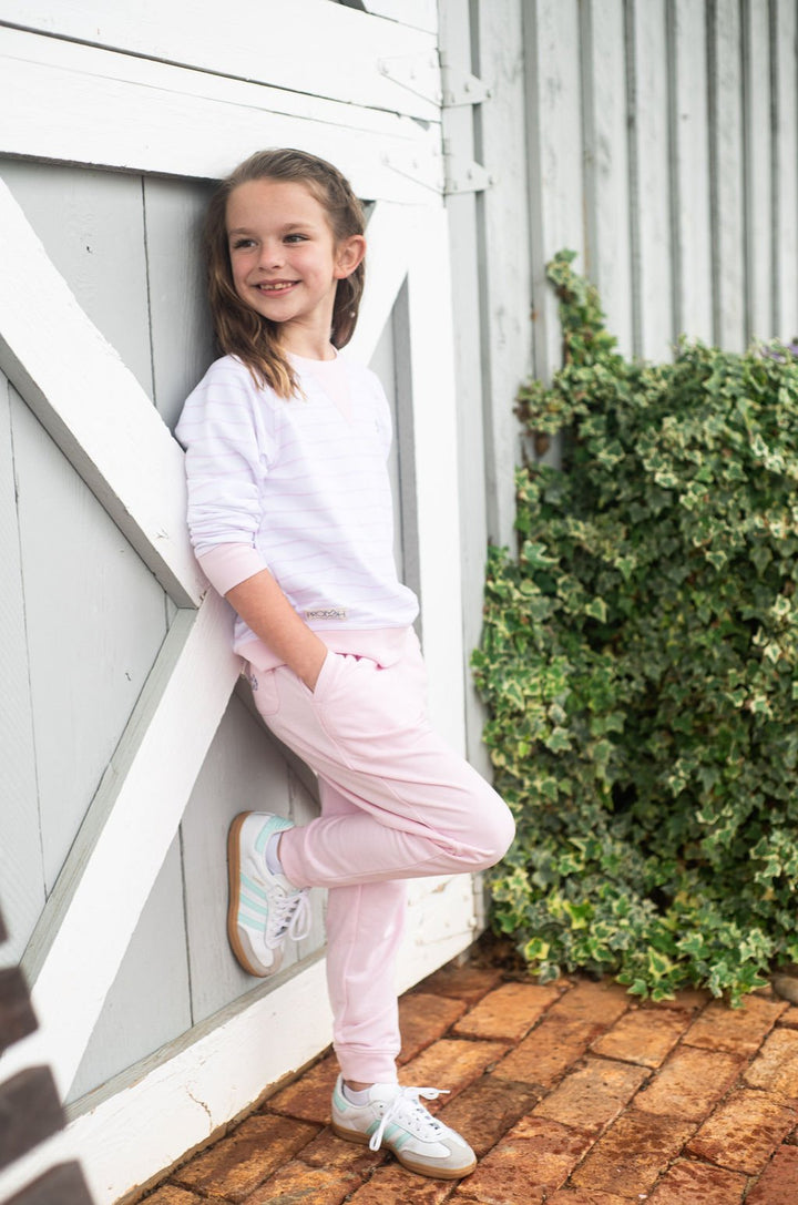 Young girl in a white shirt and pink pants standing against a white wooden wall.