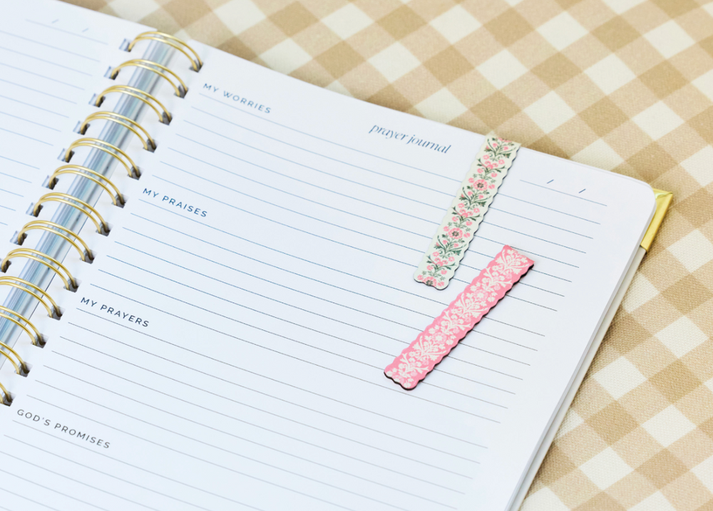 An open spiral-bound prayer journal with lined pages, decorated with two pink floral washi tape bookmarks, placed on a beige and white checkered fabric surface.