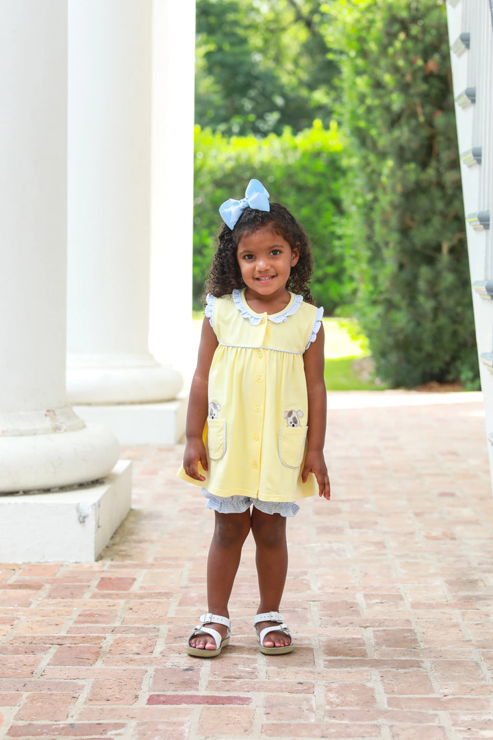 LITTLE GIRL WEARING YELLOW SLEEVELESS TOP WITH BLUE RUFFLE TRIM AND PUPPIES IN THE POCKETS.
