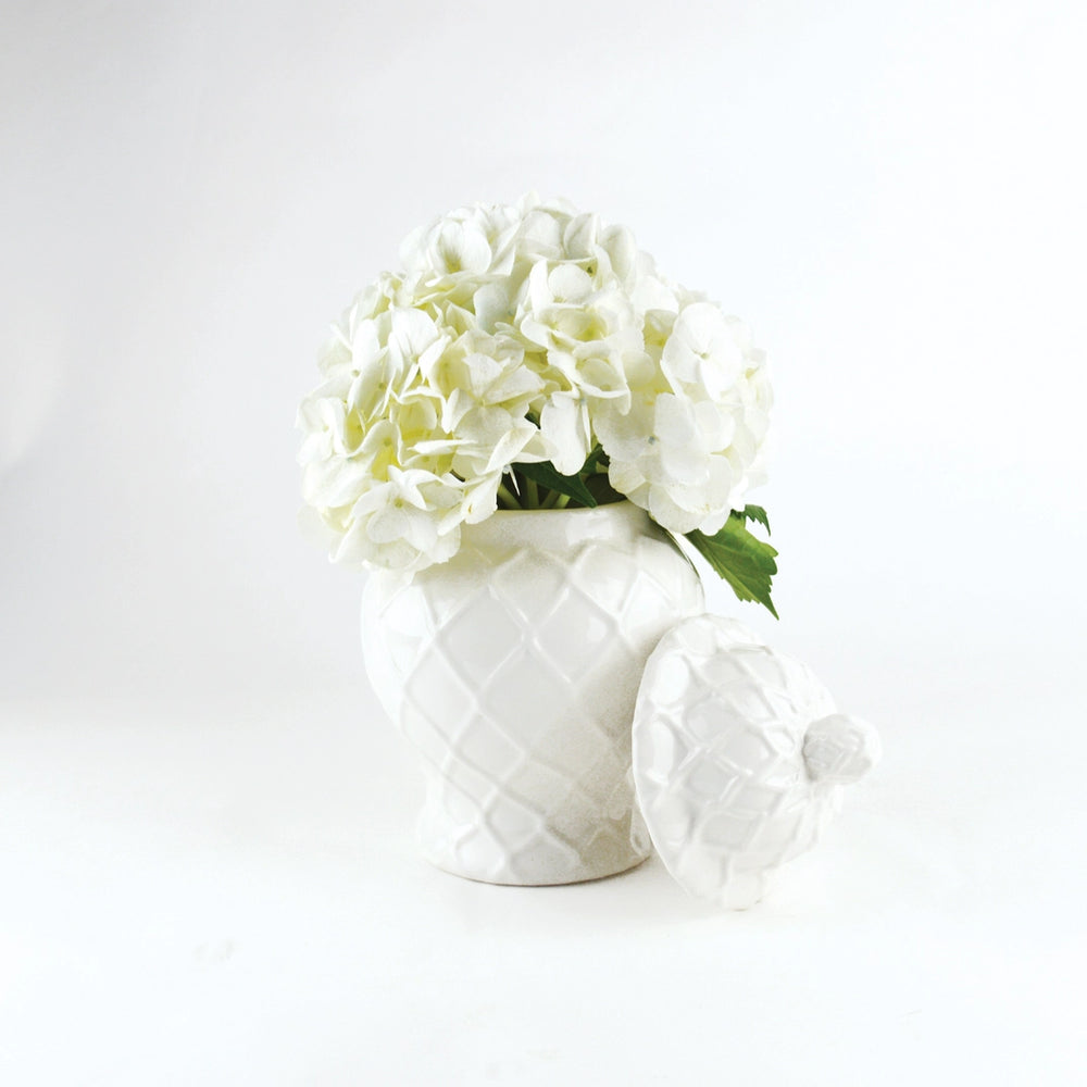 White ceramic pineapple-shaped vase with a lid containing white flowers on a light gray background
