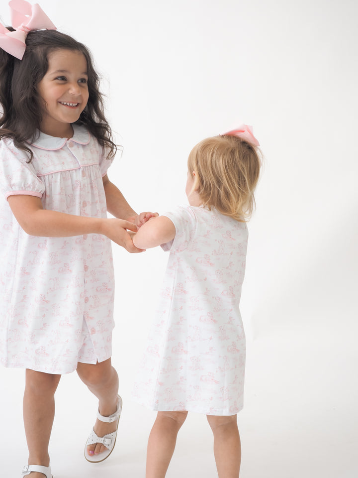Two young girls in matching white dresses with pink puppy pattern standing on a white background.