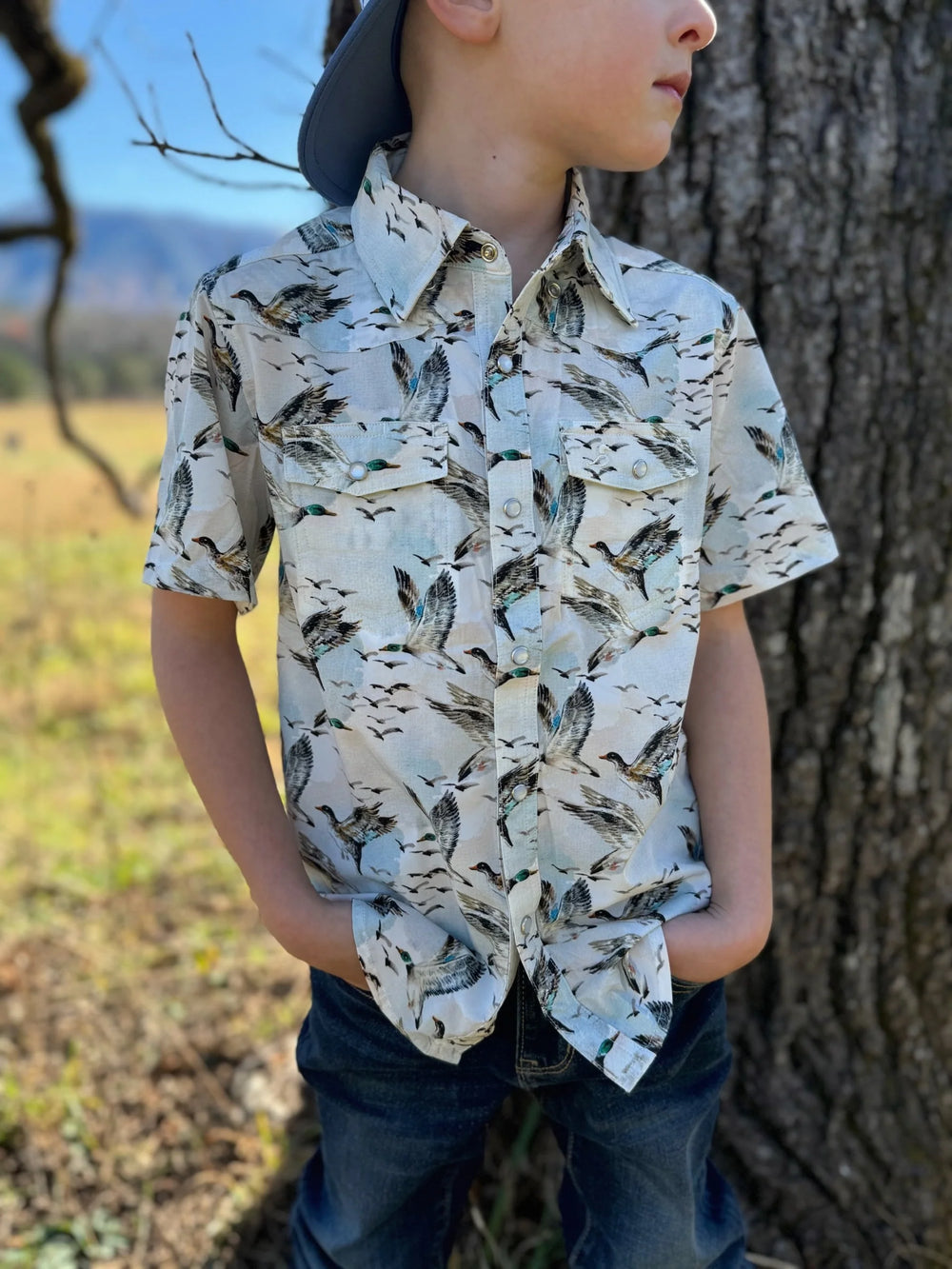 Child wearing a patterned shirt standing next to a tree outdoors