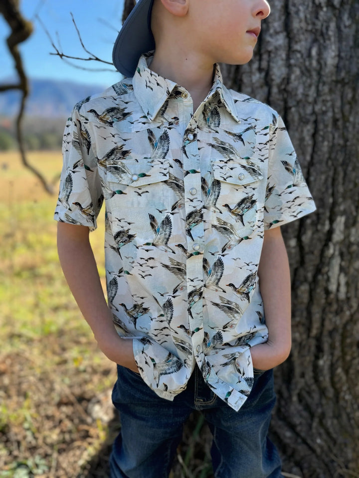 Child wearing a patterned shirt standing next to a tree outdoors