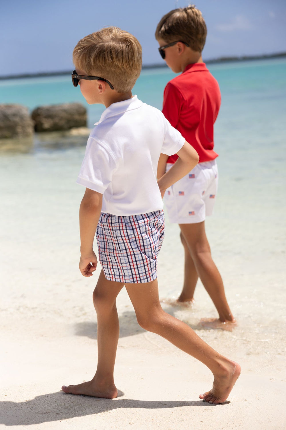 Two boys walking on a sandy beach with clear blue water and sky.