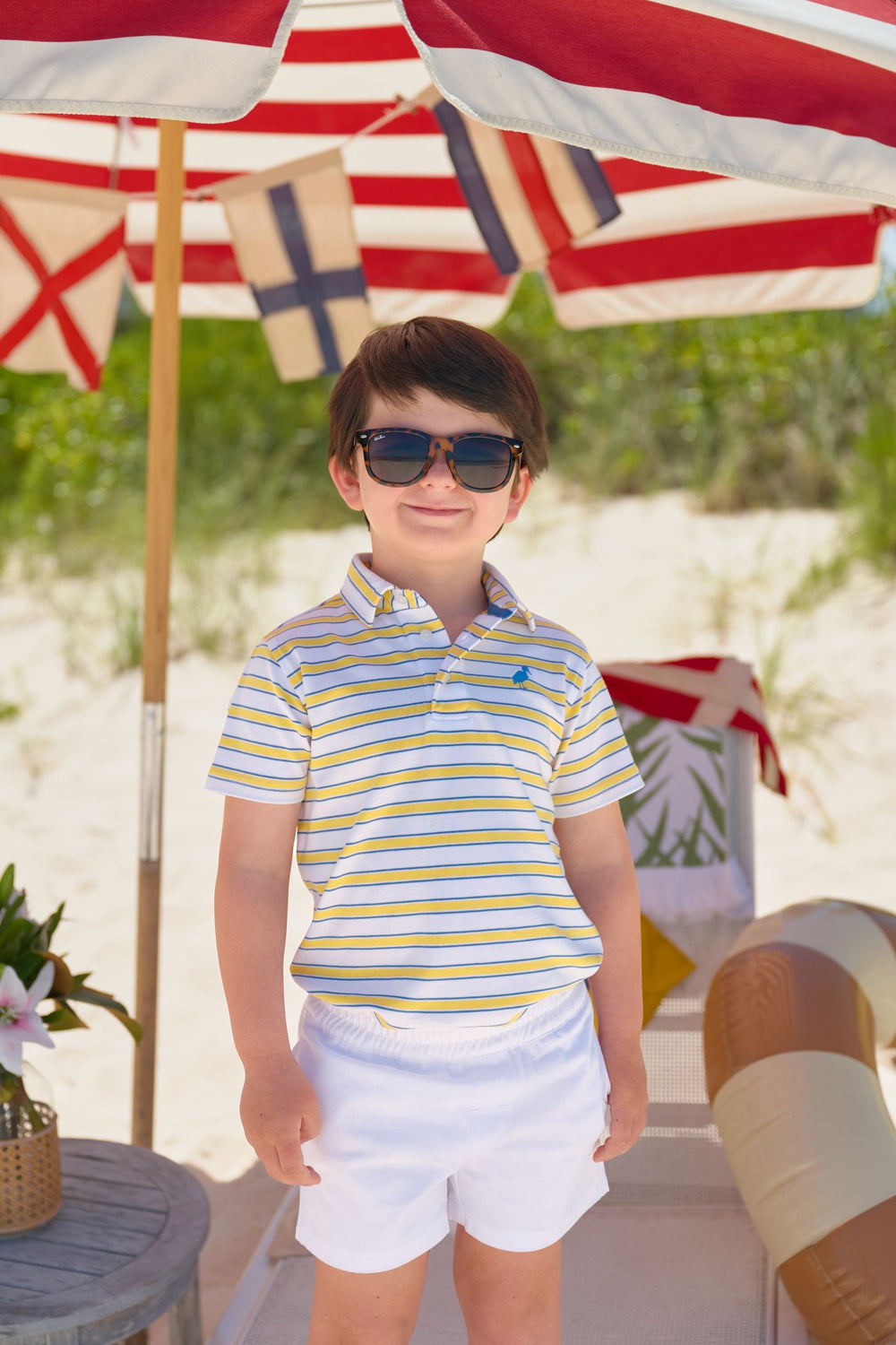 Young boy in striped polo shirt and shorts standing under a red and white umbrella with nautical flags.