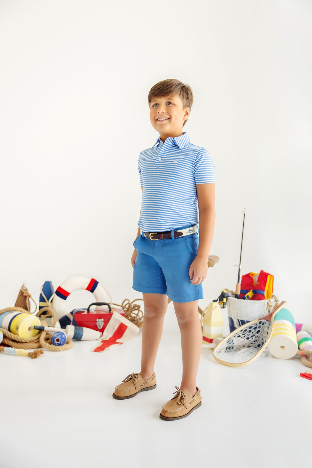 Young boy in a blue striped shirt and shorts standing in front of a white background with sports equipment.